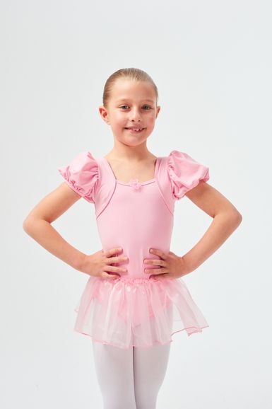 Junges Mädchen in einem rosa Ballett-Outfit und weißen Strumpfhosen posiert mit den Händen in den Hüften.