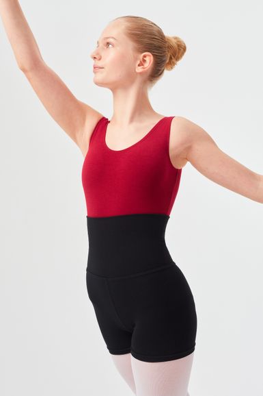 Eine junge Tänzerin in einem roten Trikot mit schwarzen Shorts posiert mit einem erhobenen Arm.