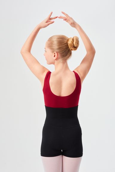 Eine Balletttänzerin in einem roten Trikot und schwarzen Shorts steht mit erhobenen Armen da und blickt von der Kamera weg.