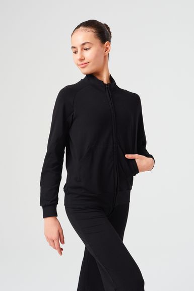 Ein Mädchen in einer schwarzen Jacke mit Reißverschluss und einer schwarzen Hose steht mit einer Hand in der Tasche.