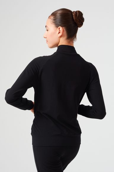 Eine Frau mit braunem Haar zu einem Dutt trägt eine schwarze Jacke mit Reißverschluss und eine schwarze Hose und blickt von der Kamera weg.