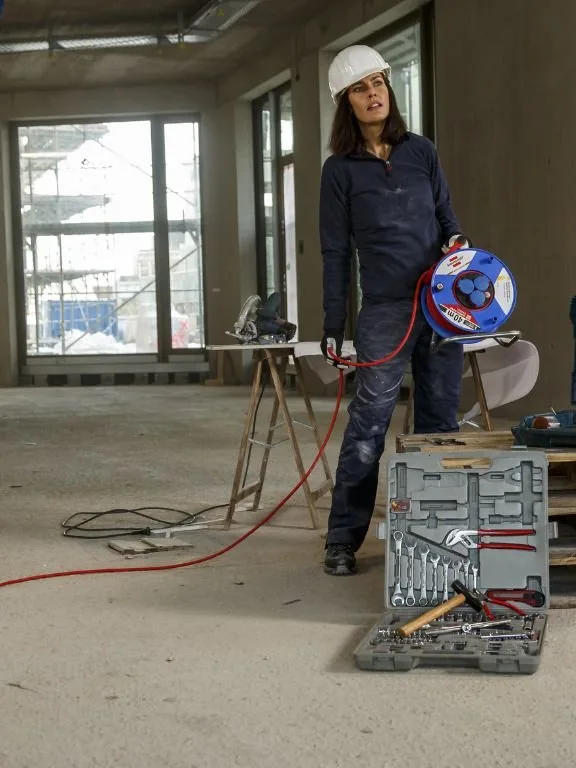 Handwerkerin mit Schutzhelm rollt auf der Baustelle das Kabel einer Brennenstuhl Kabeltrommel ab