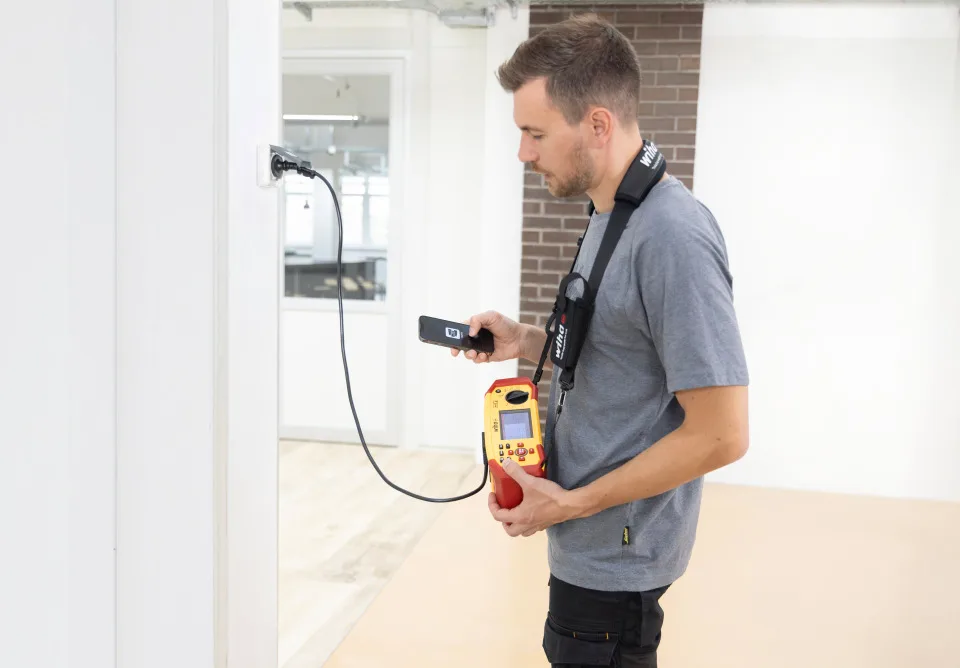 Elektriker mit dem Wiha Installationstester MFT one am Umhängegurt misst an einer Steckdose und liest das Ergebnis auf dem Smartphone ab