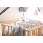 Livipur Livipur Beistellbett Ola aus Bio Erlenholz_2763 Das Livipur Beistellbett Ola aus Bio Erlenholz von Livipur mit einer grauen Decke die über die Seite drapiert ist steht in einem sanft beleuchteten Schlafzimmer mit einem Bett einem Fernseher und einer dekorativen Leuchte im Hintergrund.