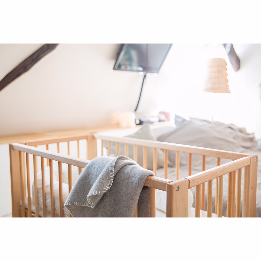 Livipur Livipur Beistellbett Ola aus Bio Erlenholz_2763 Das Livipur Beistellbett Ola aus Bio Erlenholz von Livipur mit einer grauen Decke die über die Seite drapiert ist steht in einem sanft beleuchteten Schlafzimmer mit einem Bett einem Fernseher und einer dekorativen Leuchte im Hintergrund.
