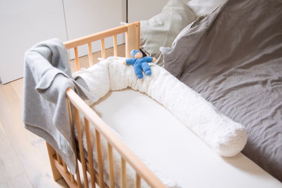 Livipur Livipur Beistellbett Ola aus Bio Erlenholz_2748 Das Livipur Beistellbett Ola aus Bio Erlenholz mit einem weichen weißen Stoßfänger einer kleinen blauen Puppe und einer grauen Decke über der Seite steht neben einem ungemachten Erwachsenenbett mit grauer Bettwäsche.