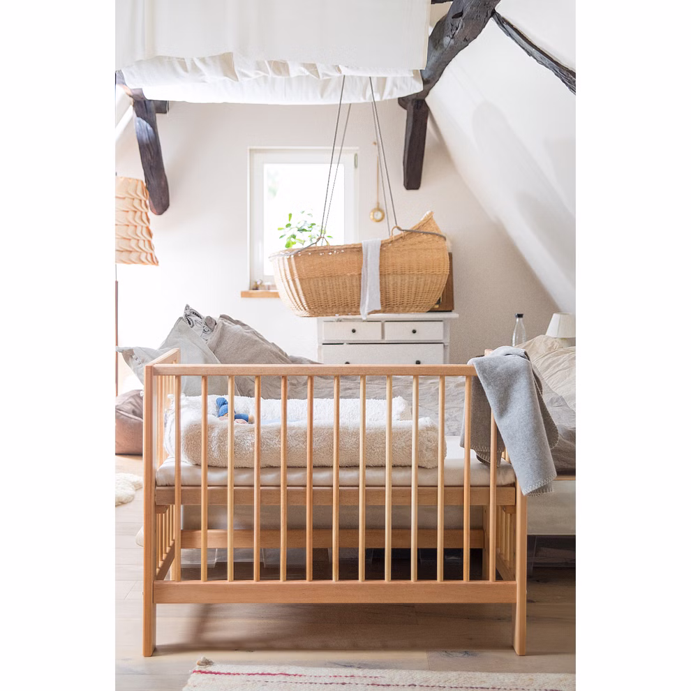 Livipur Livipur Beistellbett Ola aus Bio Erlenholz_2771 Ein gemütliches Kinderzimmer mit dem Livipur Beistellbett Ola aus Bio Erlenholz von Livipur im Vordergrund einem Sofa mit Kissen und einer hängenden Korbwiege am Fenster. Der Raum hat schräge Decken mit sichtbaren Balken und sanftes natürliches Licht.