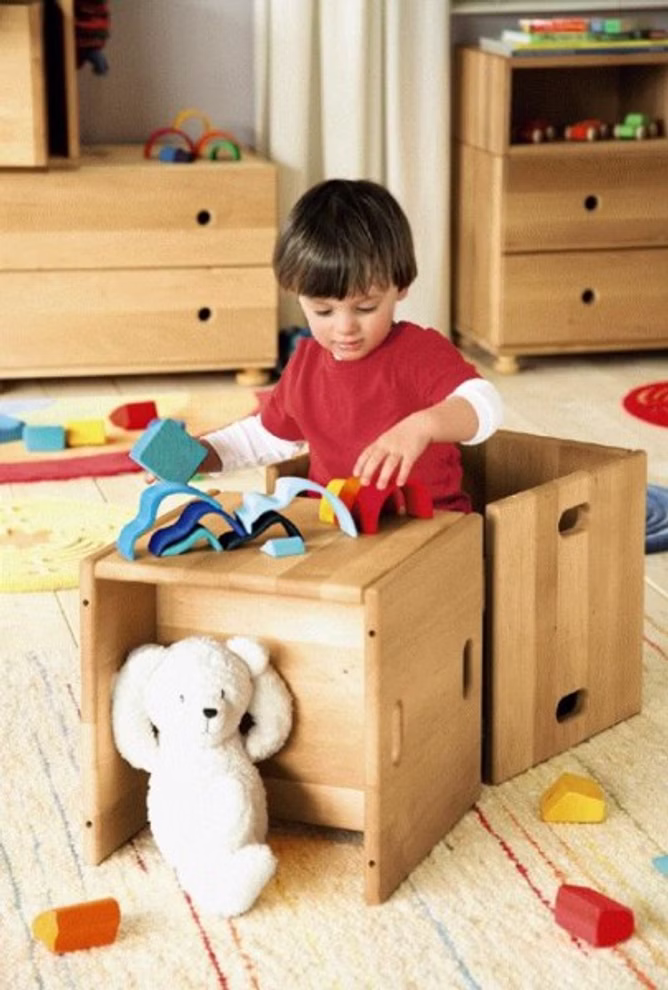 Livipur Livipur Zauberwürfel Hocker 2er Set _3514 Ein kleines Kind in einem roten Hemd spielt an einem Livipur Zauberwürfel Hocker (2er Set) Holztisch. Darunter liegt ein weißer Plüschbär während andere Spielzeuge von Livipur auf dem Teppichboden verstreut sind.