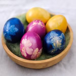 Eine mit grünem Filz ausgekleidete Holzschale enthält sechs bunte Ostereier - zwei blaue zwei gelbe und zwei violette - die mit weißen Blumen- und Blattmotiven verziert sind. Die Eier sind mit den Ostereierfarben von Auro aus rein natürlicher Basis gefärbt