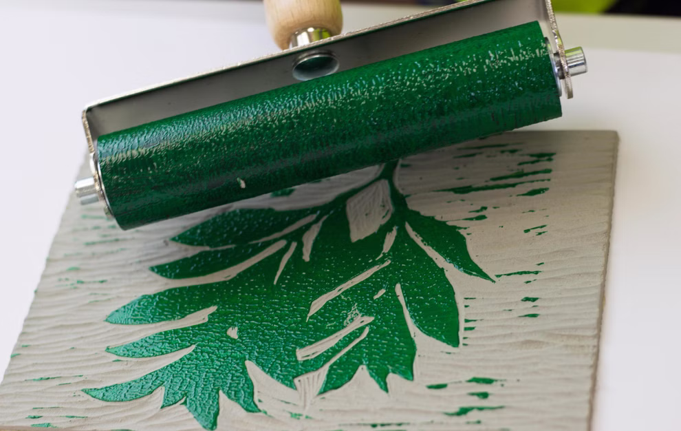 Daler-Rowney Professional Linolplatte A5 Daler Rowney Professional _14082 Eine Walze verteilt grüne Tinte auf der Daler-Rowney Professional Linolplatte A5 die mit einem Blattmotiv versehen ist und demonstriert so den Druckprozess.