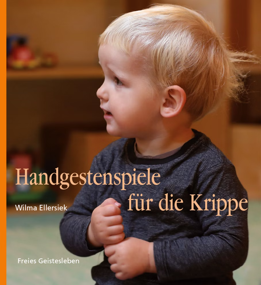 Freies Geistesleben Handgestenspiele für die Krippe Wilma Ellersiek_8060 Ein junges blondes Kind in einem dunklen langärmeligen Hemd sitzt in einem Haus. Das Buch "Handgestenspiele für die Krippe Wilma Ellersiek" (Freies Geistesleben) ist zu sehen der Hintergrund ist sanft verschwommen.