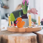 Ein Grimms Geburtstagsring klein natur Ø 22 cm mit Kerzen einem grünen Baum und einer orangefarbenen Eichhörnchenfigur sitzt auf einem Holztisch. Im Hintergrund sind eine verschwommene Person und eine rosa Hyazinthe zu sehen.