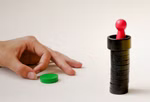 Steffen Spiele Spiel - Rettet Rapunzel! _14334 Eine Hand greift nach einer grünen Figur aus Spiel - Rettet Rapunzel! von Steffen Spiele während ein roter Bauer auf gestapelten schwarzen Spielsteinen balanciert und einen Turm auf einer weißen Fläche bildet.