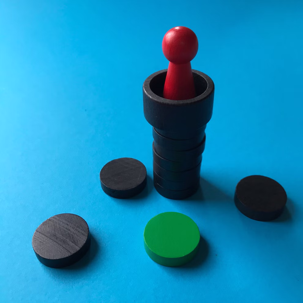 Steffen Spiele Spiel - Rettet Rapunzel! _14333 Eine rote Spielfigur aus Spiel - Rettet Rapunzel! von Steffen Spiele steht auf gestapelten schwarzen Scheiben auf einer blauen Fläche daneben liegen drei schwarze Scheiben und eine grüne Scheibe.