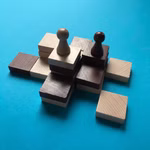 Zwei Holzfiguren aus Spiel - Volterra von Steffen Spiele eine helle und eine dunkle stehen auf gestapelten Holzklötzen in verschiedenen Farbtönen angeordnet in einem Raster auf einem leuchtend blauen Hintergrund.