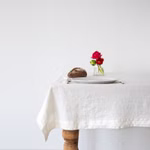 Linen Tales Tischdecke aus Leinen weiß 100 % Leinen_15284 140x300 Auf einem Holztisch der mit Tales Tischdecke aus Leinen weiß 100 % Leinen drapiert ist stehen ein Teller Besteck ein kleiner Laib Brot und eine Glasvase mit roten und rosa Blumen vor einer schlichten weißen Wand. 140x300
