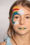 Ein junges Mädchen mit braunen Haaren trägt Regenbogenschminke mit Wolken Glitzern und einer Sonne die mit dem Schminkstift von Namaki Cosmetics in 12 Farben hergestellt wurde. Ihre Lippen sind blau und gelb. Sie ist hellblau gekleidet und blickt in die Ka fuchsia