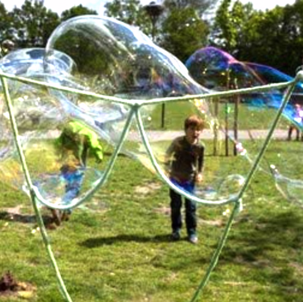 BubbelLab Riesenseifenblasen Set für Feste_32229 Ein Kind auf einer Wiese im Freien benutzt das BubbelLab Riesenseifenblasen Set für Feste um riesige Seifenblasen zu erzeugen wobei im Hintergrund Bäume und ein Weg zu sehen sind.