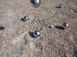Schildkröt Boule Set aus Stahl 2 x 3 Spielkugeln Zielkugel Abstandsmesser_18043 Das Boule-Set aus Stahl von Schildkröt (2 x 3 Spielkugeln Zielkugel Abstandsmesser) liegt auf Kies wobei eine Stahlkugel in der Luft über der hölzernen Zielkugel liegt und einen Schatten auf den Boden wirft.
