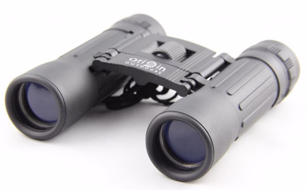 Origin Outdoors Fernglas 10-fache Vergrößerung_18199 Ein kompaktes schwarzes Fernglas 10-fache Vergrößerung mit blau getönten Gläsern ruht auf einer weißen Fläche und trägt den Markennamen Origin Outdoors auf dem Steg zwischen den Okularen.