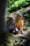 Freies Geistesleben Fingerpüppchen häkeln und nähen Jana Tröger_18596 Zwei lächelnde Kinder kauern an einem Baum in einem sonnenbeschienenen Wald und halten kleine handgefertigte Puppen aus "Fingerpüppchen häkeln und nähen" von Jana Tröger Freies Geistesleben umgeben von grünem Laub und gedämpftem Sonnenlicht.