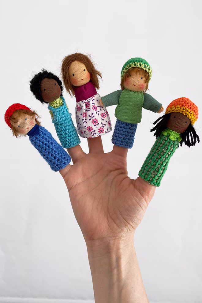 Freies Geistesleben Fingerpüppchen häkeln und nähen Jana Tröger_18593 Fünf Fingerpuppen aus "Fingerpüppchen häkeln und nähen Jana Tröger" von Freies Geistesleben sind auf einer Hand abgebildet jede in bunten gestrickten Kleidern und Mützen mit Garnhaaren und gestickten Gesichtern vor einem schlichten weißen Hintergrund.