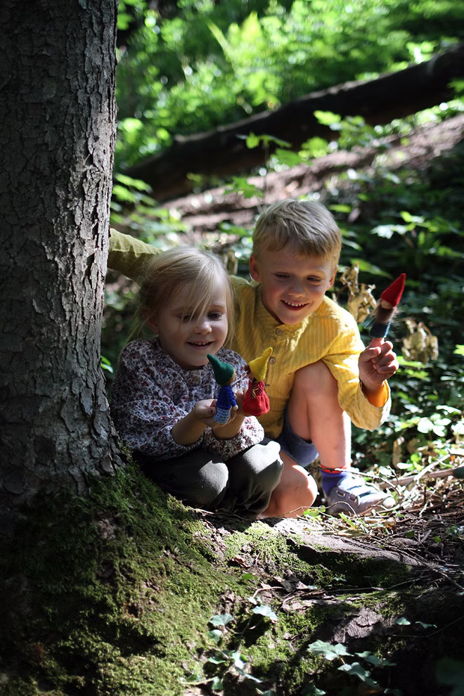 Freies Geistesleben Fingerpüppchen häkeln und nähen Jana Tröger_18596 Zwei lächelnde Kinder kauern an einem Baum in einem sonnenbeschienenen Wald und halten kleine handgefertigte Puppen aus "Fingerpüppchen häkeln und nähen" von Jana Tröger Freies Geistesleben umgeben von grünem Laub und gedämpftem Sonnenlicht.
