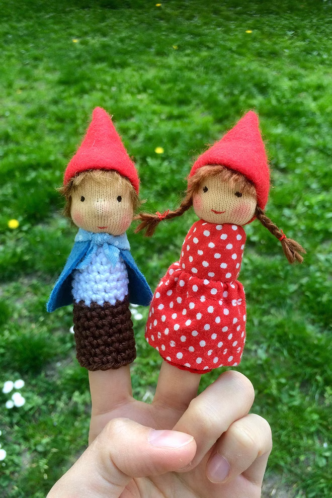 Freies Geistesleben Fingerpüppchen häkeln und nähen Jana Tröger_18598 Zwei Fingerpuppen "Fingerpüppchen häkeln und nähen Jana Tröger" von Freies Geistesleben - eine in Blau und Braun mit rotem Hut die andere mit rotem gepunktetem Kleid und Hut - werden auf Fingern vor einer grasbewachsenen Kulisse im Freien gezeigt.