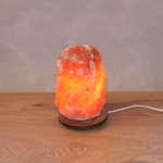Eine Himalaya Salt Dreams Salzkristalllampe mit Holzsockel (ca. 15-2 kg) steht auf einer hölzernen Oberfläche und leuchtet warm mit dem sichtbaren weißen Stromkabel im Hintergrund.
