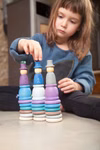 Grapat Grapat Holzspielzeug Winter Set _19624 Ein kleines Kind sitzt auf dem Boden und stapelt konzentriert bunte Holzringe aus dem Grapat Holzspielzeug Winter Set auf drei Steckpuppen in Blau Lila und Weiß von Grapat.