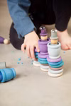 Grapat Grapat Holzspielzeug Winter Set _19625 Ein Kind stapelt bunte Grapat Holzspielzeug Winter Set Holzringe und Becher von Grapat zu Türmen auf einem hellen Boden mit verstreuten blauen Teilen und teilweise sichtbaren nackten Füßen.