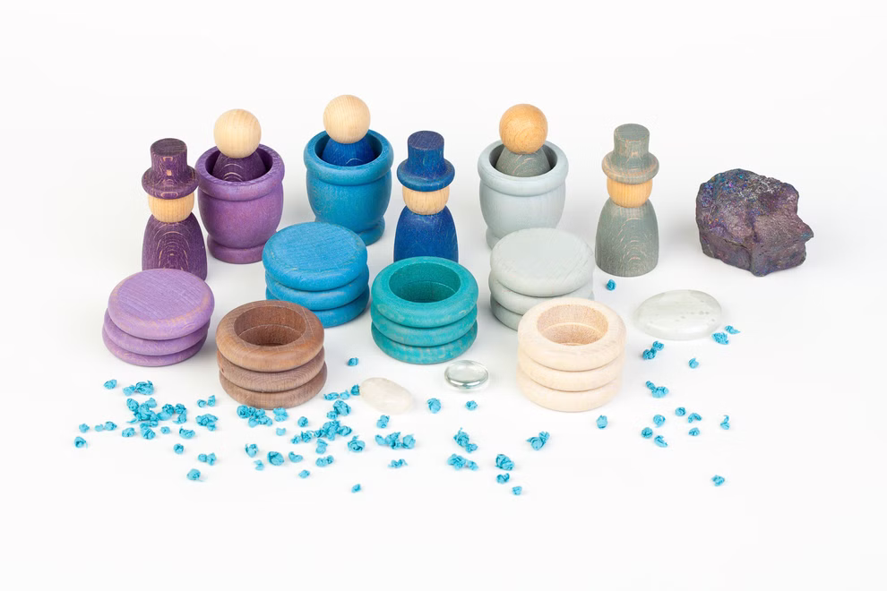 Grapat Grapat Holzspielzeug Winter Set _19623 Das Grapat Holzspielzeug Winter Set von Grapat besteht aus farbenfrohen Holzpuppen und Bechern in lila blauen und blaugrünen Tönen die auf einem weißen Hintergrund mit Steinen einem Felsen und blauen Kieselsteinen im Vordergrund stehen.