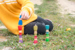 Grapat Grapat Holzspielzeug Zauberer _19640 Ein Kind in einem gelben Pullover sitzt im Gras und stapelt Grapat Holzspielzeug Zauberer von Grapat zu Türmen. Bunte Holzbecher und Wäscheklammern umgeben sie kleine Blumen sind verstreut und eine Klammer ist mit einer winzigen Blume gekrönt.
