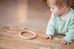 Ein Kleinkind in einem hellgrünen Hemd lehnt an einem Holztisch und starrt auf das Grapat Holzspielzeug 3 Ringe groß Ø 13 cm von Grapat das auf dem Tisch steht. Der sanft verschwommene Hintergrund hebt das Spielzeug hervor.