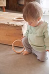 Ein kleines Kind in einem mintgrünen Hemd und einer grauen Hose kniet auf dem Boden und hält einen Grapat Holzspielzeug 3 Ringe groß Ø 13 cm Holzring von Grapat mit einer rustikalen Holzkiste und einem weiteren Ring in der Nähe.