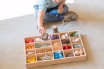 Ein Kind sitzt auf dem Boden und spielt mit der Grapat Sortierbox Holz Setzkasten von Grapat. Es nimmt einen Stein aus dem hölzernen Sortierkasten der mit bunten Objekten wie Steinen Holzformen Tannenzapfen und Muscheln gefüllt ist.