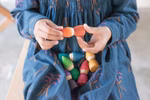 Ein Kind in einem blauen Kleid sitzt und spielt mit Grapat Holzspielzeug Regenbogen Zwerge von Grapat wobei es einige Puppen in der Hand hält und weitere bunte Puppen auf dem Schoß hat.