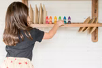 Ein kleines Kind mit langen braunen Haaren arrangiert Grapat Holzspielzeug Regenbogen Zwerge von Grapat auf einem Holzregal vor einer weißen strukturierten Wand.