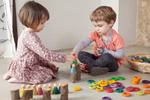 Zwei kleine Kinder sitzen auf dem Boden und spielen mit der Grapat Große Holzbox von Grapat. Sie ordnen Holzfiguren an und stapeln Ringe aus der Box. Ein Korb in der Nähe enthält weitere Spielsachen während sie sich auf ihre kreative Tätigkeit konzentriere