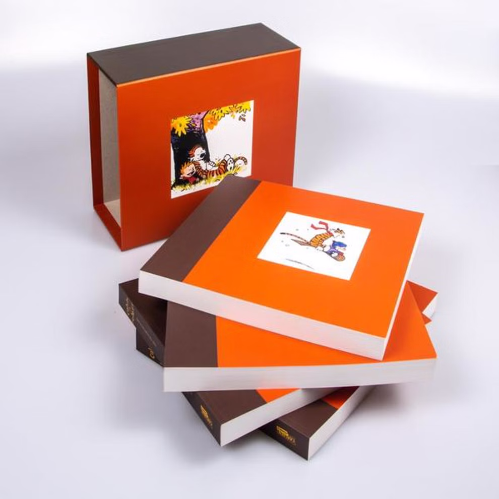 Carlsen Verlag Calvin und Hobbes Gesamtausgabe - Paperback Bill Watterson_19700 Drei Taschenbücher der "Calvin und Hobbes Gesamtausgabe" von Bill Watterson (Carlsen Verlag) mit Cartoon-Einband sind neben einem passenden Schuber mit einem Cartoon-Bild auf weißem Hintergrund gestapelt.