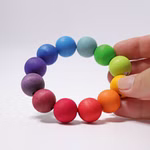 Eine Hand hält den Grimm's Regenbogen Perlenring (Ø 9 cm) von Grimm's mit großen runden Holzperlen (Ø 2 cm) in aufeinanderfolgenden Regenbogenfarben vor einem schlichten weißen Hintergrund.