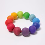 Der Grimm's Regenbogen Perlenring (Ø 9 cm) von Grimm's besteht aus großen runden 2 cm großen Perlen in einem Regenbogen-Farbverlauf die kreisförmig auf weißem Grund angeordnet sind.