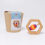 Grimm's Sechseck Rassel Ø 8 cm eine sechseckige Holzrassel mit bunten Ringen wird neben ihrer Kartonverpackung gezeigt die auf der Vorderseite die Marke Grimm's und Angaben zum Spielzeug enthält.