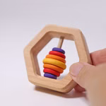 Eine Hand hält Grimm's Sechseck Rassel (Ø 8 cm) von Grimm's eine sechseckige Babyrassel aus Holz mit einem zentralen Stab und sieben bunten Ringen in Rot Orange Gelb und Lila. Der Hintergrund ist einfarbig weiß.