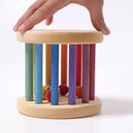 Eine Hand berührt den oberen Teil des Grimm's Regenbogen Babyrollers Ø 15 cm von Grimm's einem Holzspielzeugzylinder mit bunten vertikalen Stäben und kleinen Kugeln im Inneren vor einem weißen Hintergrund.