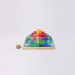 Eine kleine Schnecke steht neben Grimms Kleiner Stufenpyramide von Grimm's - einer bunten Pyramide aus rechteckigen Holzklötzen die in einem Farbverlauf auf einem quadratischen Tablett angeordnet sind - vor einem schlichten weißen Hintergrund.