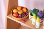 Ein ERZI Kaufladen Apfel aus Holz steht in einer Holzschale mit Spielzeugkirschen auf einem Regal daneben eine Kiste mit Spielzeuggurken und -auberginen und im Vordergrund stehen zwei kleine Flaschen mit gelben Verschlüssen.