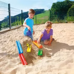 Zwei kleine Kinder spielen in einem Sandkasten mit den ERZI Wasser- und Murmelstraße 5 Spuren und benutzen bunte Rampen um Wasser zu schütten und Rinnen zu bilden. Im Hintergrund sind grüne Hügel Bäume und ein Drahtzaun zu sehen.