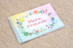 Das Meine Freunde Freundebuch von Mummel & Hummel Malwerkstatt ist ein spiralgebundenes Notizbuch mit einem pastellfarbenen geblümten Einband und rotem deutschem Text der auf beigem Stoff liegt.