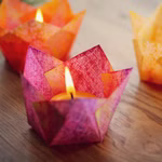 Drei lebhafte lotusförmige Origami-Halter zeigen jeweils eine brennende Kerze der Karl Schubert Gemeinschaft Bienenwachs Teelicht Ø 38 cm auf einer hölzernen Oberfläche die der Szene strukturierte Farbe und Wärme verleiht. 1